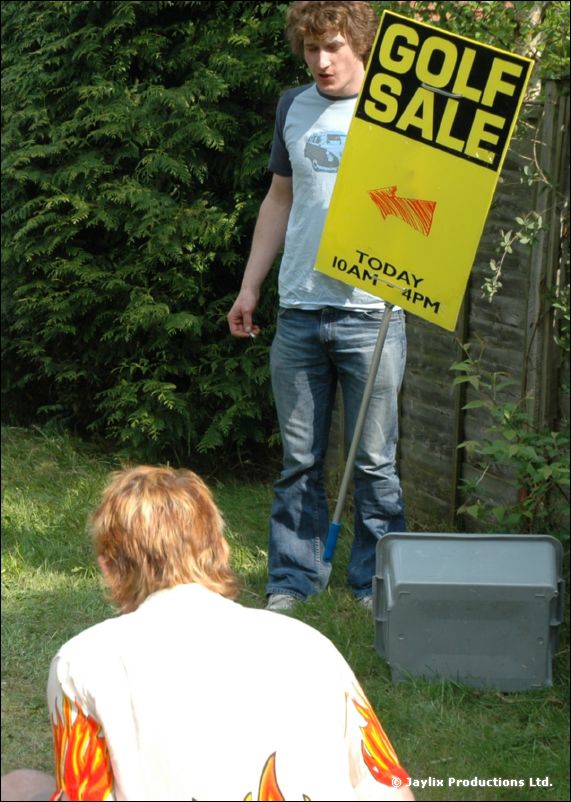 A man in a blue top holds a Golf Sale sign in a garden, with a person in a flame shirt in front