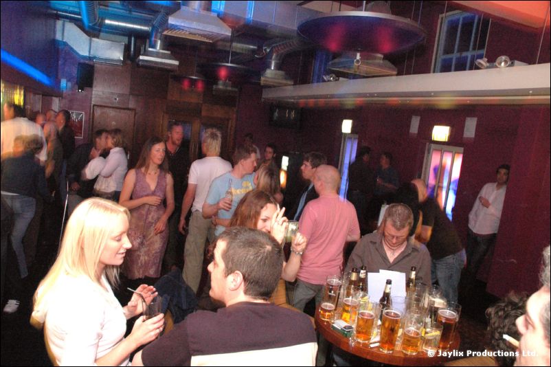 People sit and stand around a table of drinks in a bar, a blonde woman in the foreground