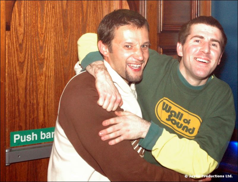 Two men hug and smile beside a wooden door with a Push bar sign, one in a Wall of Sound top