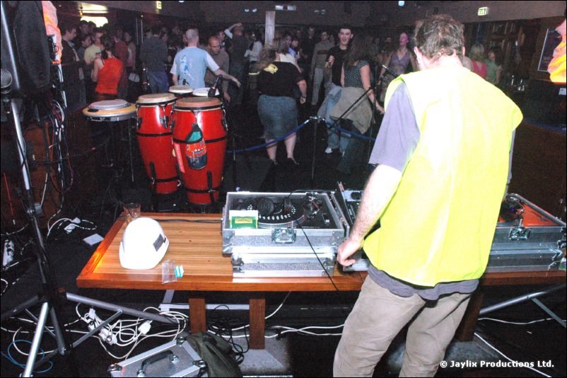 A person in a yellow hi-vis vest stands at DJ decks on a table beside conga drums, with dancers behind
