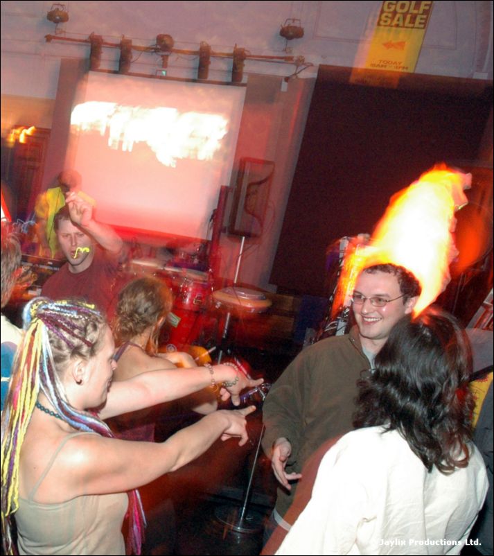 A dancer with rainbow braids points towards the camera near a flame effect, with a screen behind
