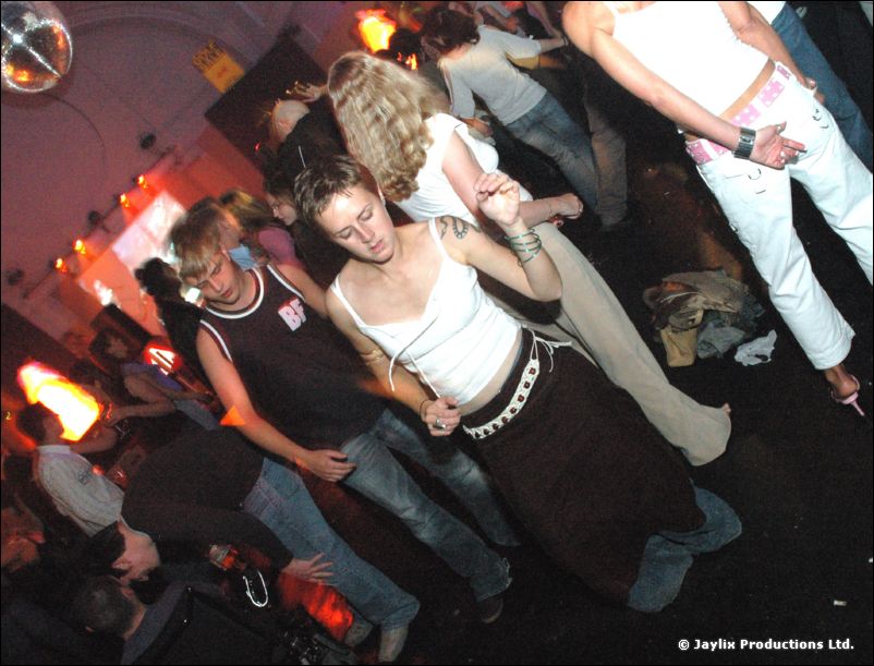 Dancers move on a floor under a mirror ball, a woman in a white top and dark skirt in the centre