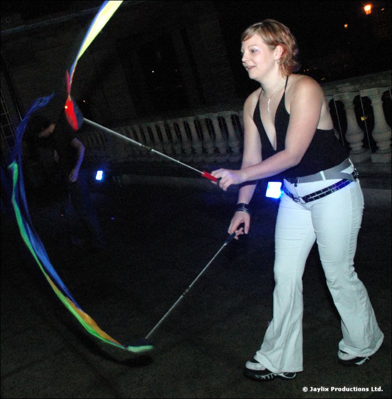 A person in a black top and white trousers swings glowing rainbow poi on a stone terrace at night