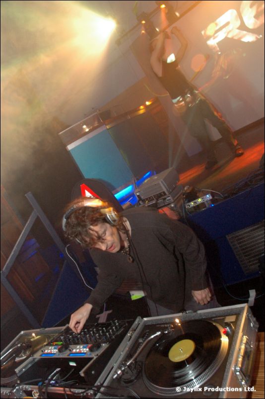 A DJ with headphones bends over turntables in a hazy, smoke-filled room