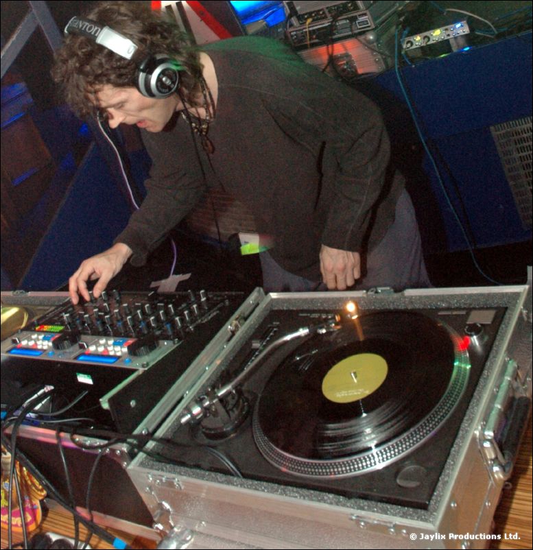 A curly-haired DJ with headphones works at turntables and a mixer