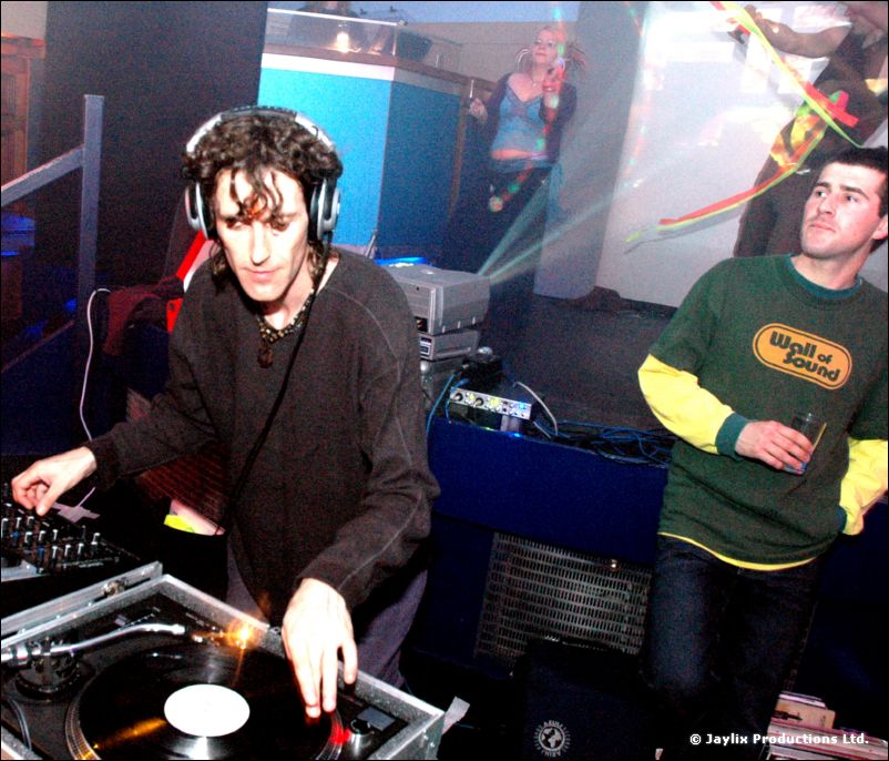A curly-haired DJ with headphones at turntables, with a man in a Wall of Sound top beside him