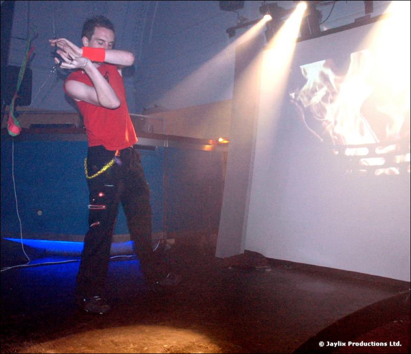 A person in a red top and black trousers dances with poi on a stage in front of a projected screen