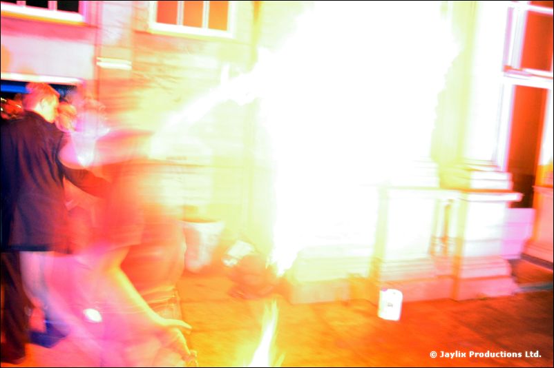A bright burst of flame outdoors beside a stone building, with blurred figures