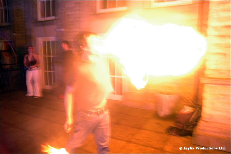 A person blows a large plume of fire outdoors beside a stone building