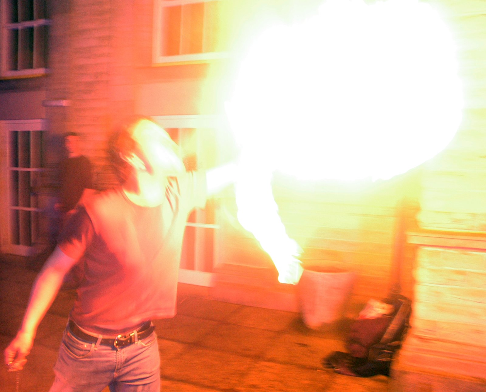 A person in a dark top blows a large plume of fire beside a stone building