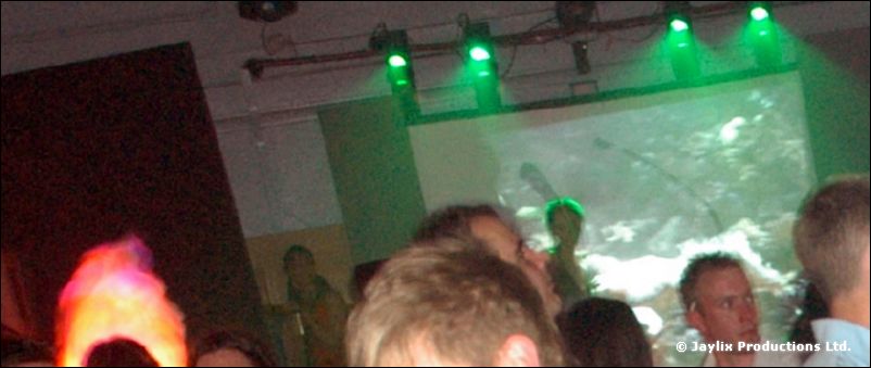A crowd faces a projected screen under green lights, with a flame effect glowing at the left