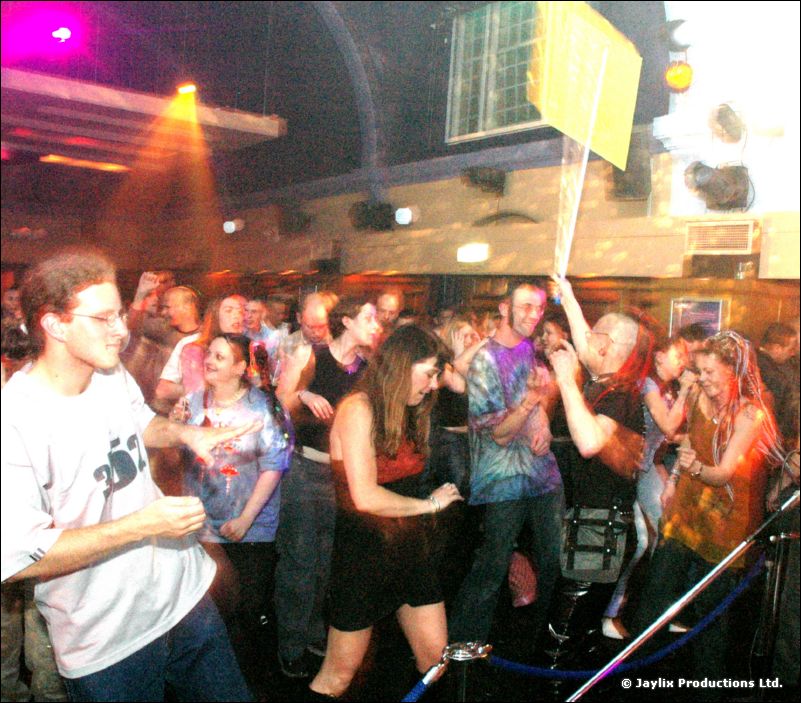 A crowd dances in a large room with arched windows, with a yellow sign held up above them
