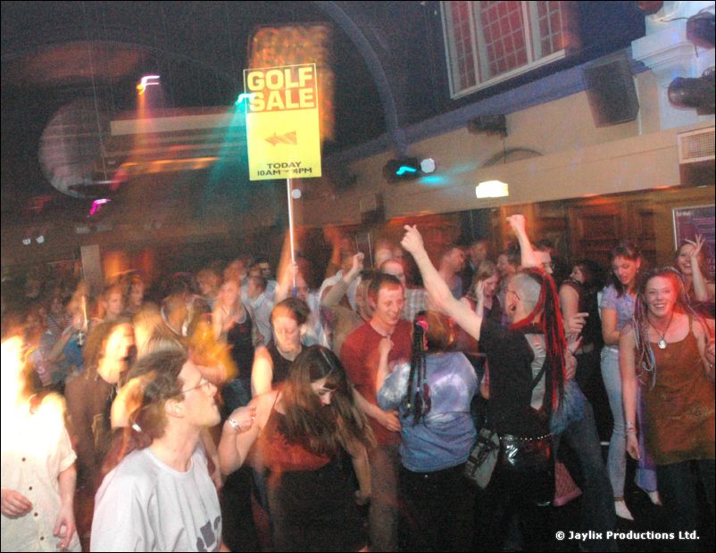 A crowd dances in a large room while someone holds up a yellow Golf Sale sign