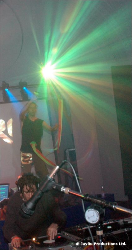 A person stands behind a DJ at decks with a microphone, with green light rays beyond