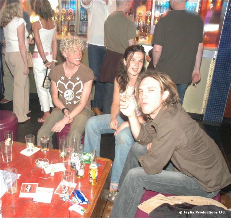 Three people sit close at a red table with drinks and flyers, one with curly blond hair