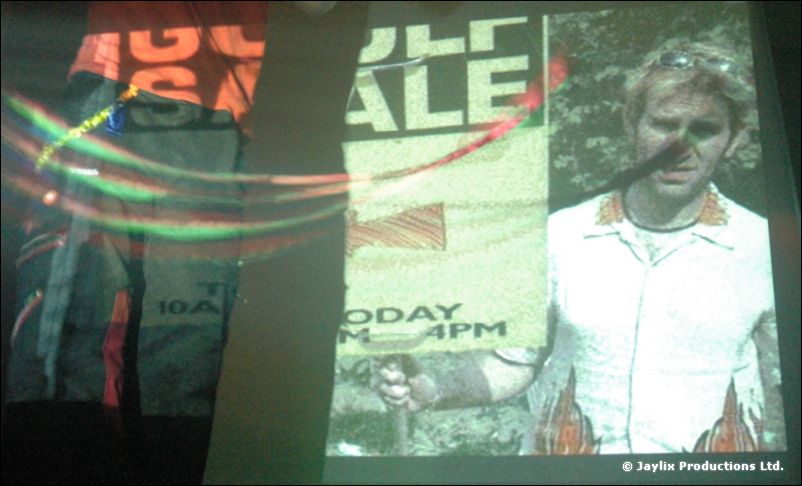 A projected image of a man in a white shirt holding a microphone, with a Golf Sale sign behind