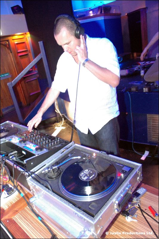 A DJ in a white shirt and headphones mixes at turntables on a wooden table
