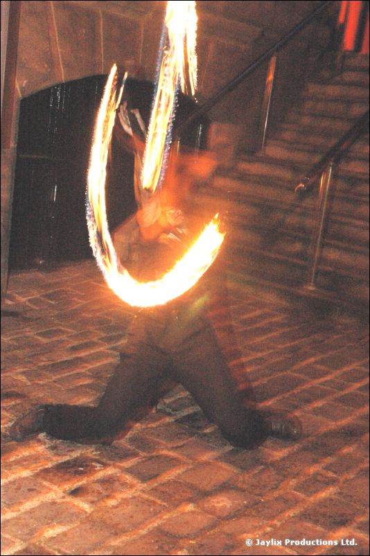 A person kneels on cobbles with a fire staff leaving trails of flame beside a stairway at night