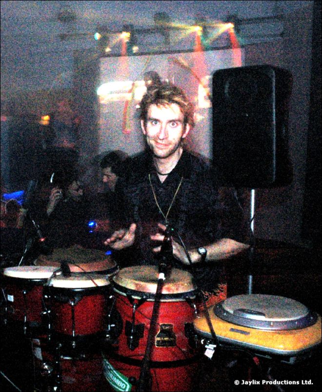 A smiling man in a dark top plays congas and drums in front of a projected screen