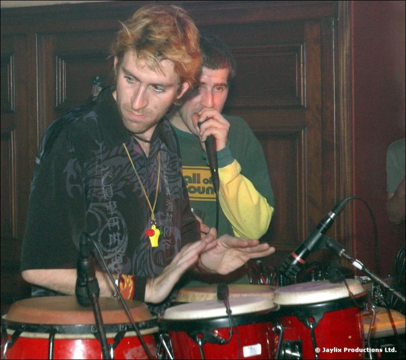 A man plays congas while another man in a Wall of Sound top speaks into a microphone behind him