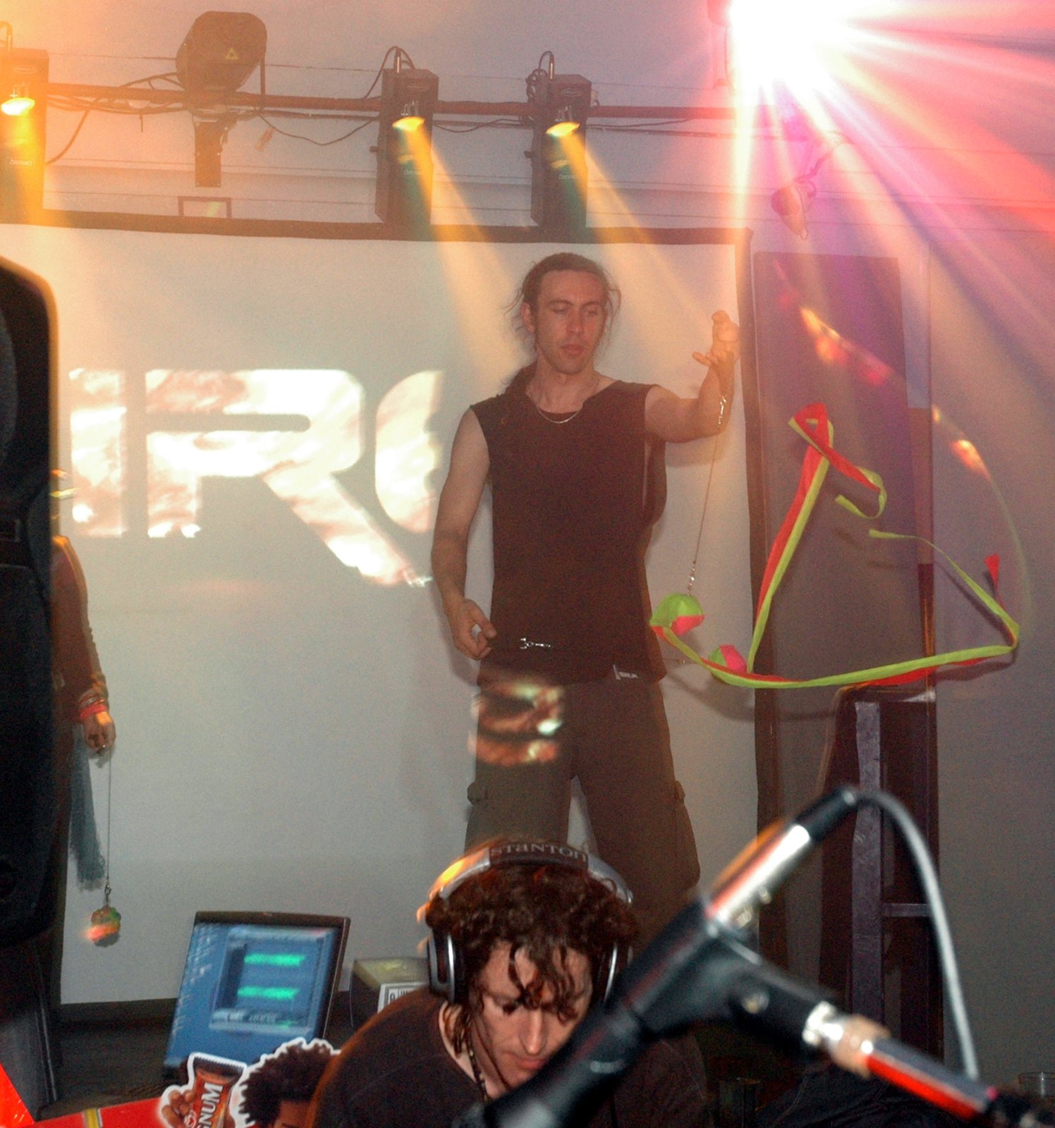 A person in a sleeveless top holds glowing poi on a stage before a screen, with a DJ in headphones below