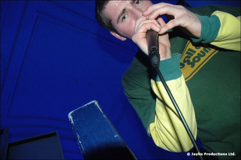 A man in a Wall of Sound top holds a microphone to his mouth against a blue wall