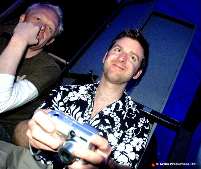 A man in a floral shirt holds a compact camera beside a man resting his hand on his face