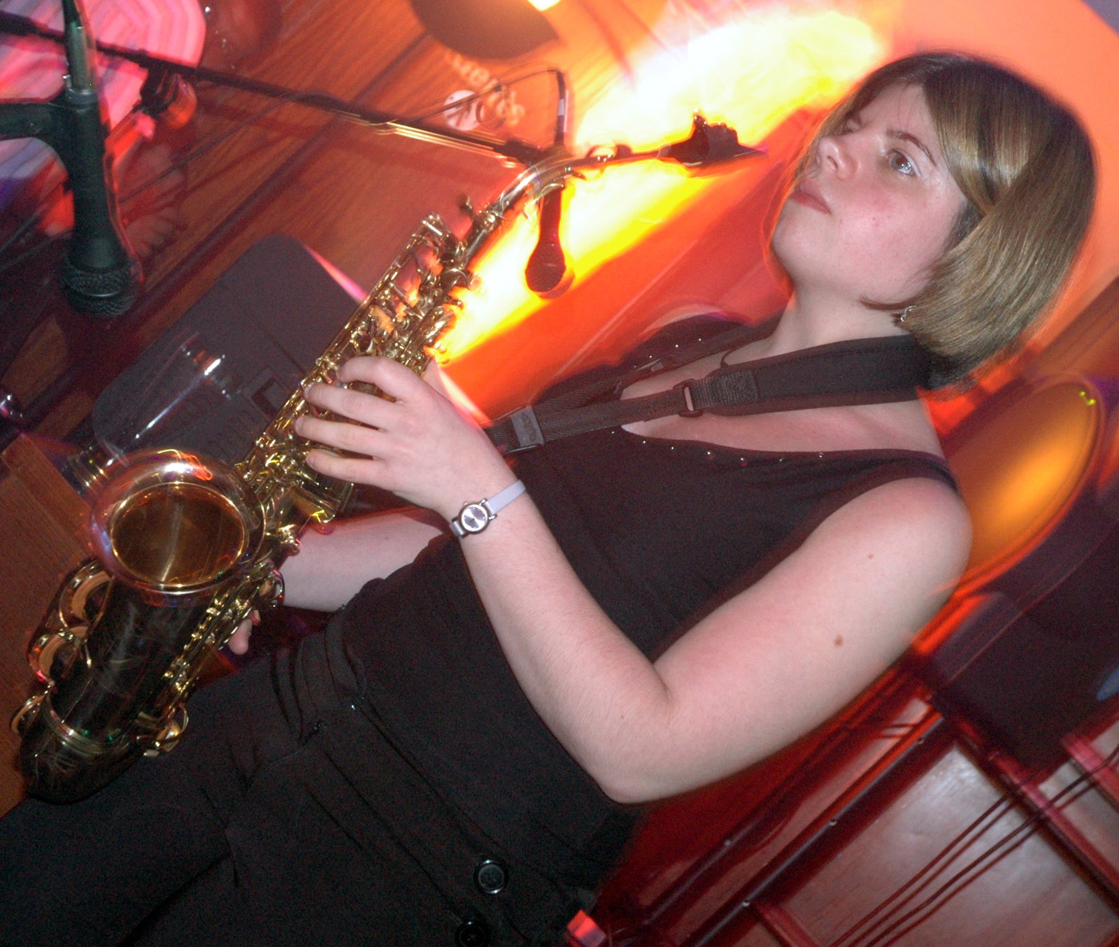 A person in a black top plays a saxophone beside a microphone stand under orange light
