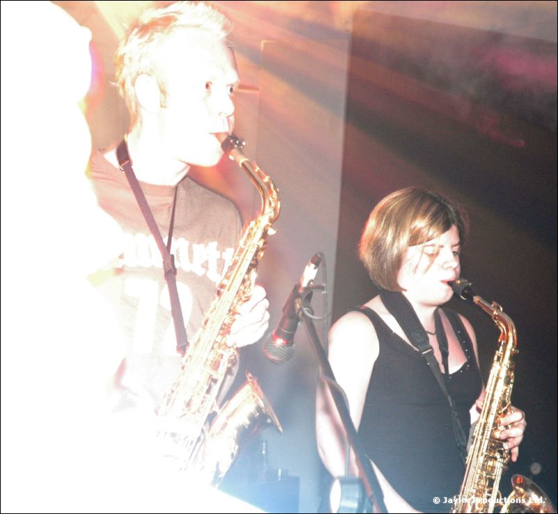Two saxophone players perform on stage, one in a black sleeveless top, in a bright washed-out flash