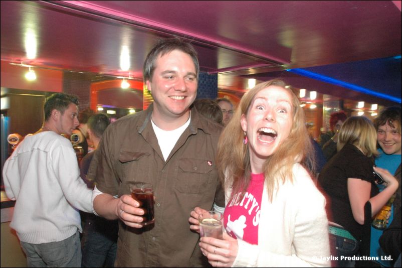 A smiling man in an olive shirt and a woman in a pink top with her mouth open hold drinks in a busy bar