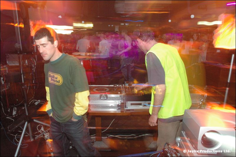 Two people in a green top and a yellow hi-vis vest stand at DJ decks in a blurred, orange-lit room
