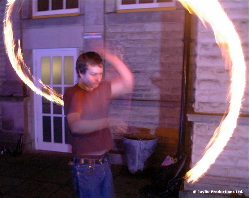 A person in a red-brown top spins two arcs of fire outside a stone building at night