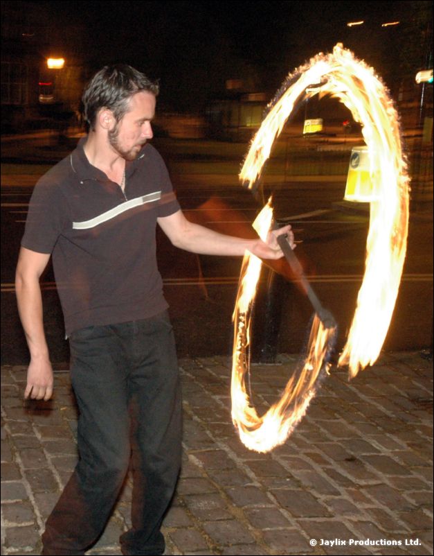 A man in a dark polo shirt swings a ring of fire on a cobbled street at night
