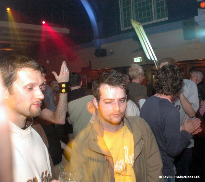 Two men in a crowded bar, one in a white top and one in a khaki jacket over a yellow t-shirt