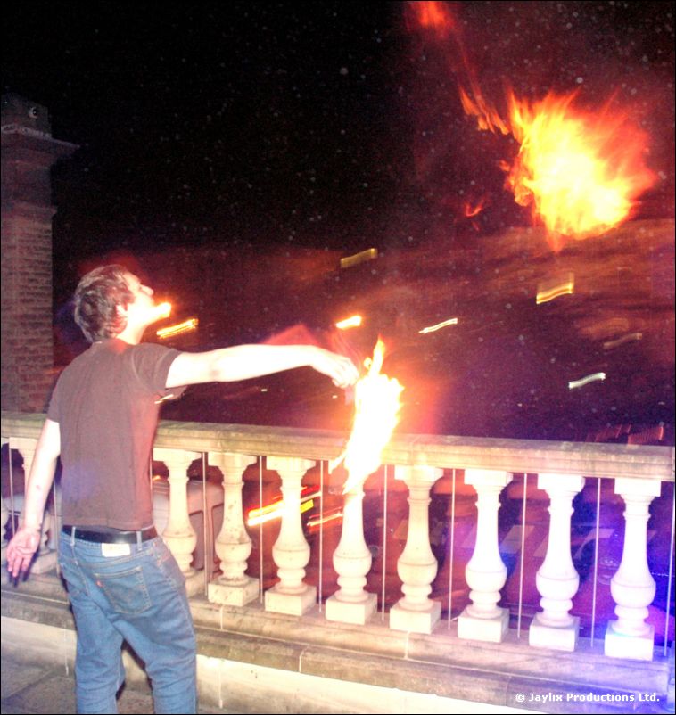 A person in a brown t-shirt swings a flaming prop beside a stone balustrade at night