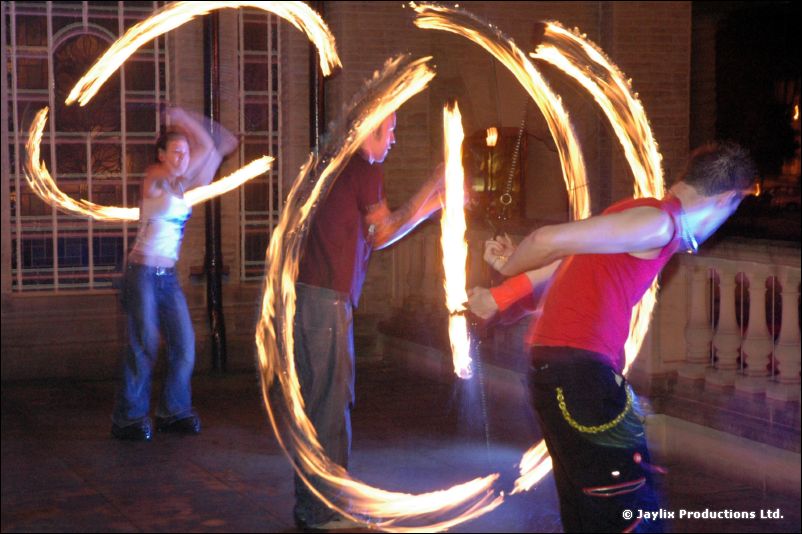 Three people spin fire on a terrace at night, one in a red sleeveless top numbered 77