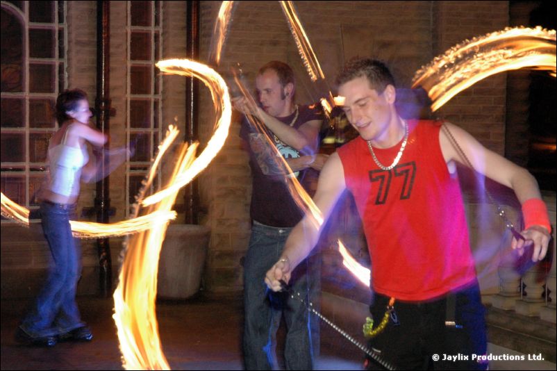 Fire performers on a terrace at night, a person in a red 77 top in the foreground with trails of fire