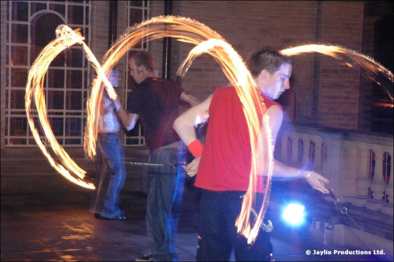 Fire performers spin flaming props on a terrace at night, one in a red sleeveless top