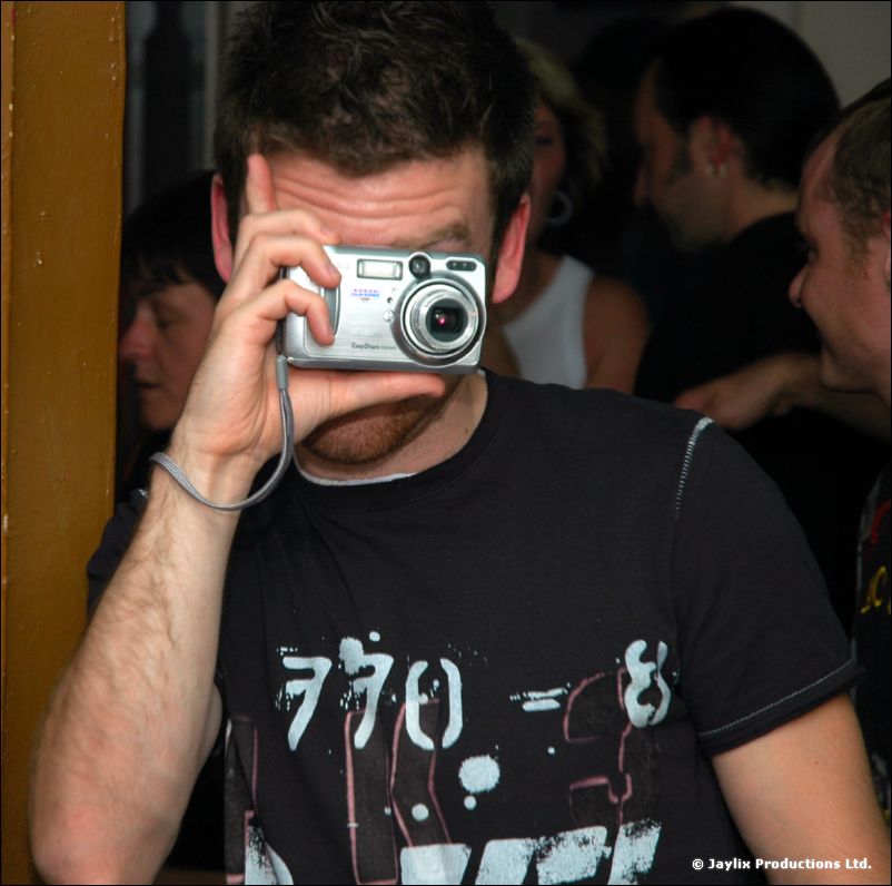 A man in a black t-shirt holds a silver compact camera to his face in a crowded room
