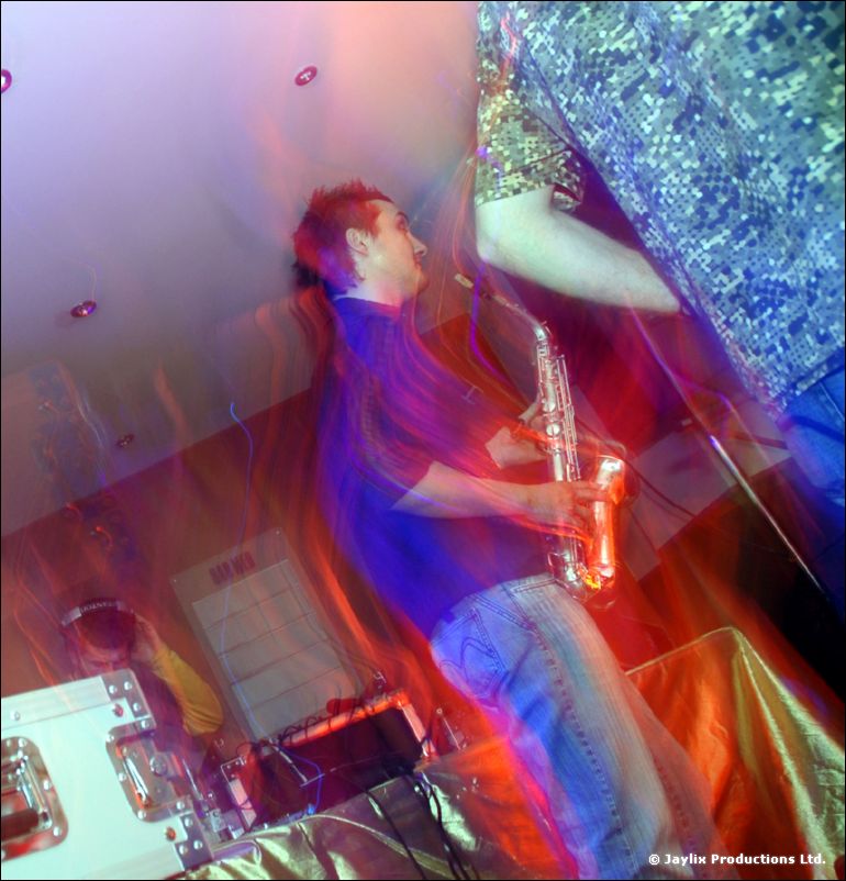 A person in a blue shirt plays a saxophone against a wall, blurred with motion and colour