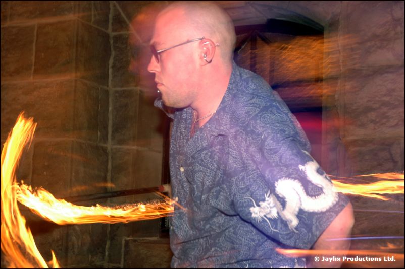 A bald man with glasses in a blue dragon-print shirt, with streaks of fire either side, outside a stone building