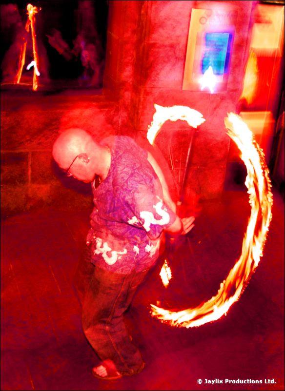 A bald man in a patterned shirt spins two trails of fire in a red-lit street at night