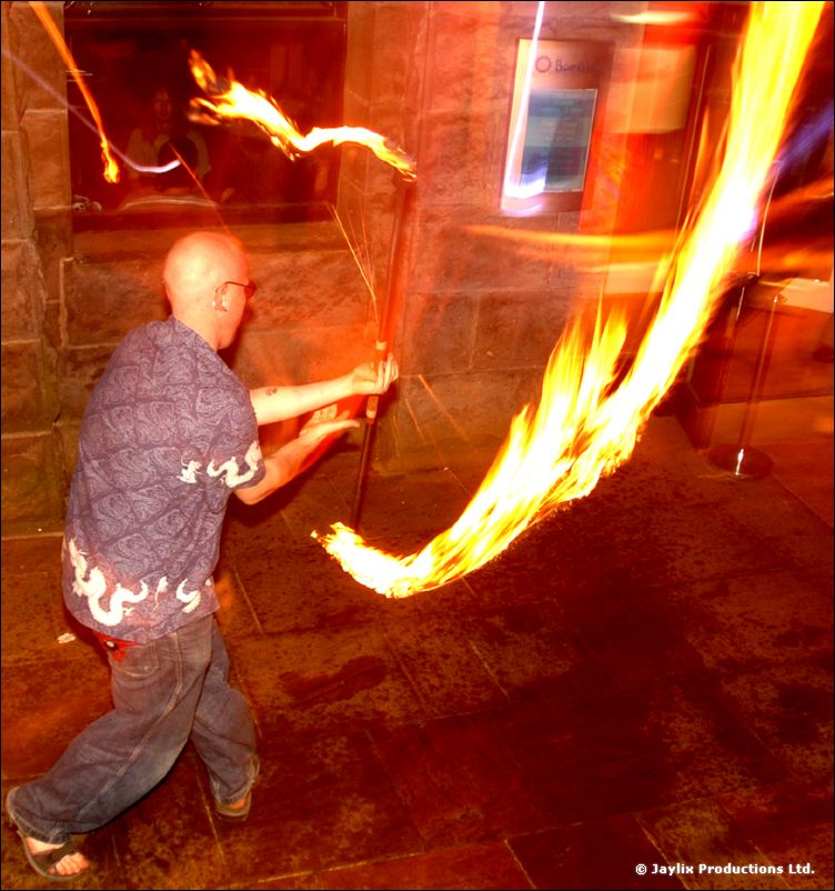 A bald man swings a flaming staff outside a stone building at night, leaving a long trail of fire