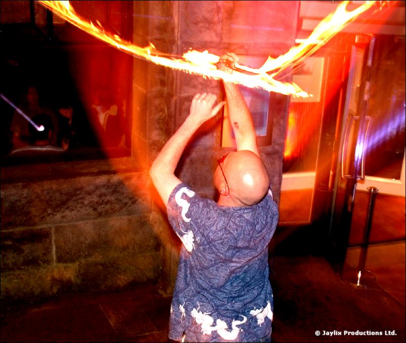 A bald man in a dragon-print shirt raises a flaming staff above his head in a red-lit street