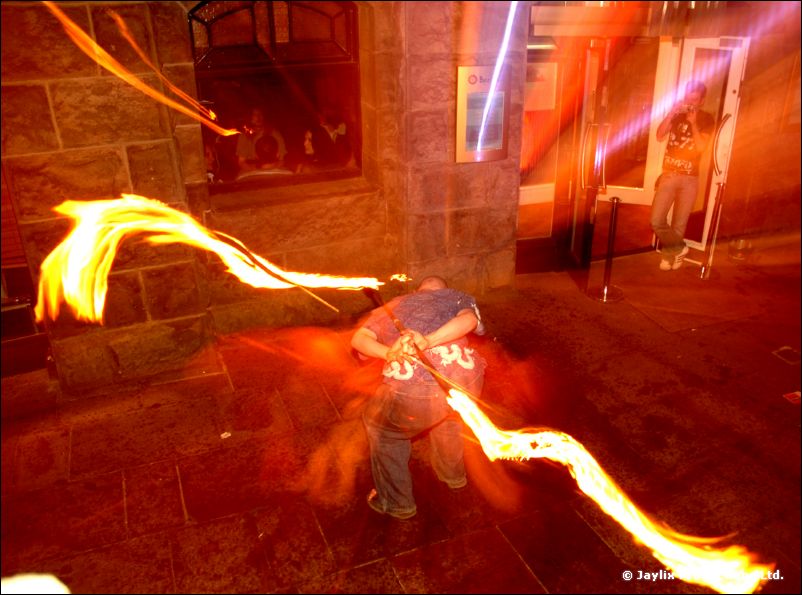 A person in a blue top spins long trails of fire in a dim stone-walled street