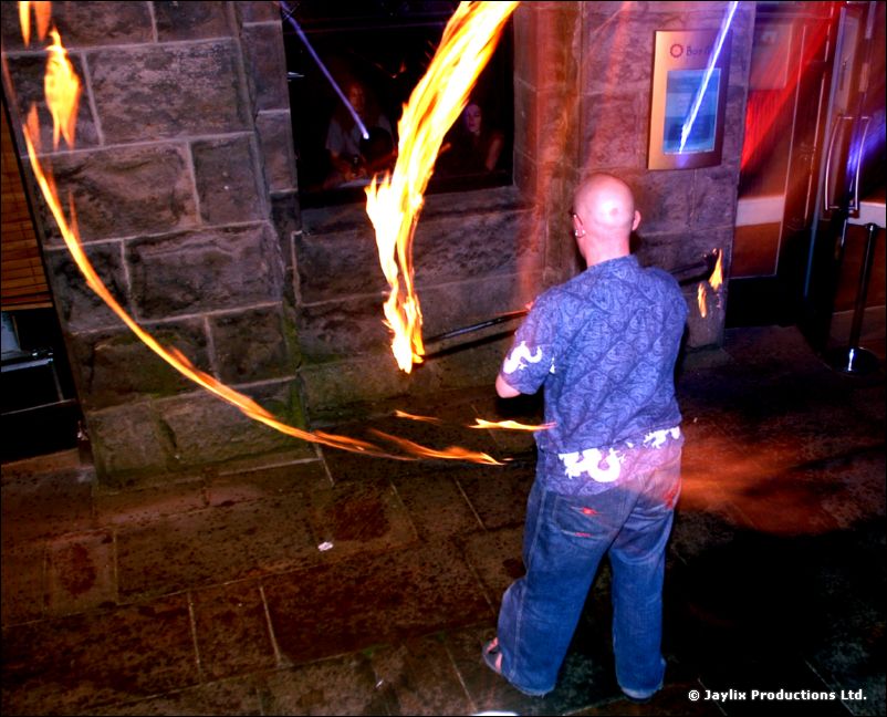 A bald man in a blue shirt stands with streaks of fire around him by a stone wall at night