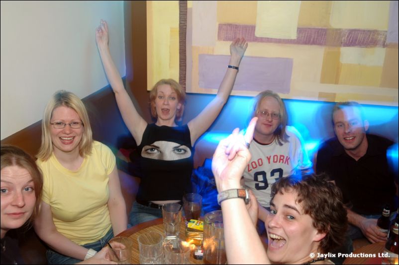 A group sits on a sofa in a bar, one person with arms raised, others laughing and pulling faces
