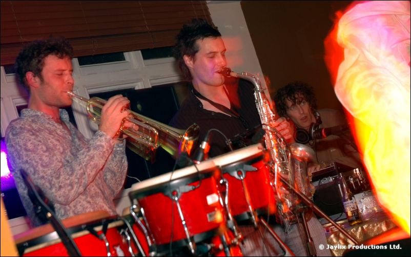 A trumpeter and a saxophonist play beside red bongo drums in a bar