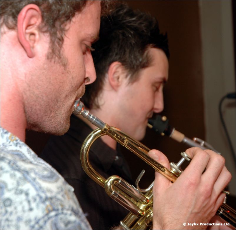 Close view of two men, one in the foreground playing a trumpet