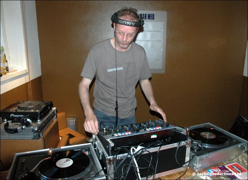 A man in headphones and a grey t-shirt stands at turntables and a mixer in a small room